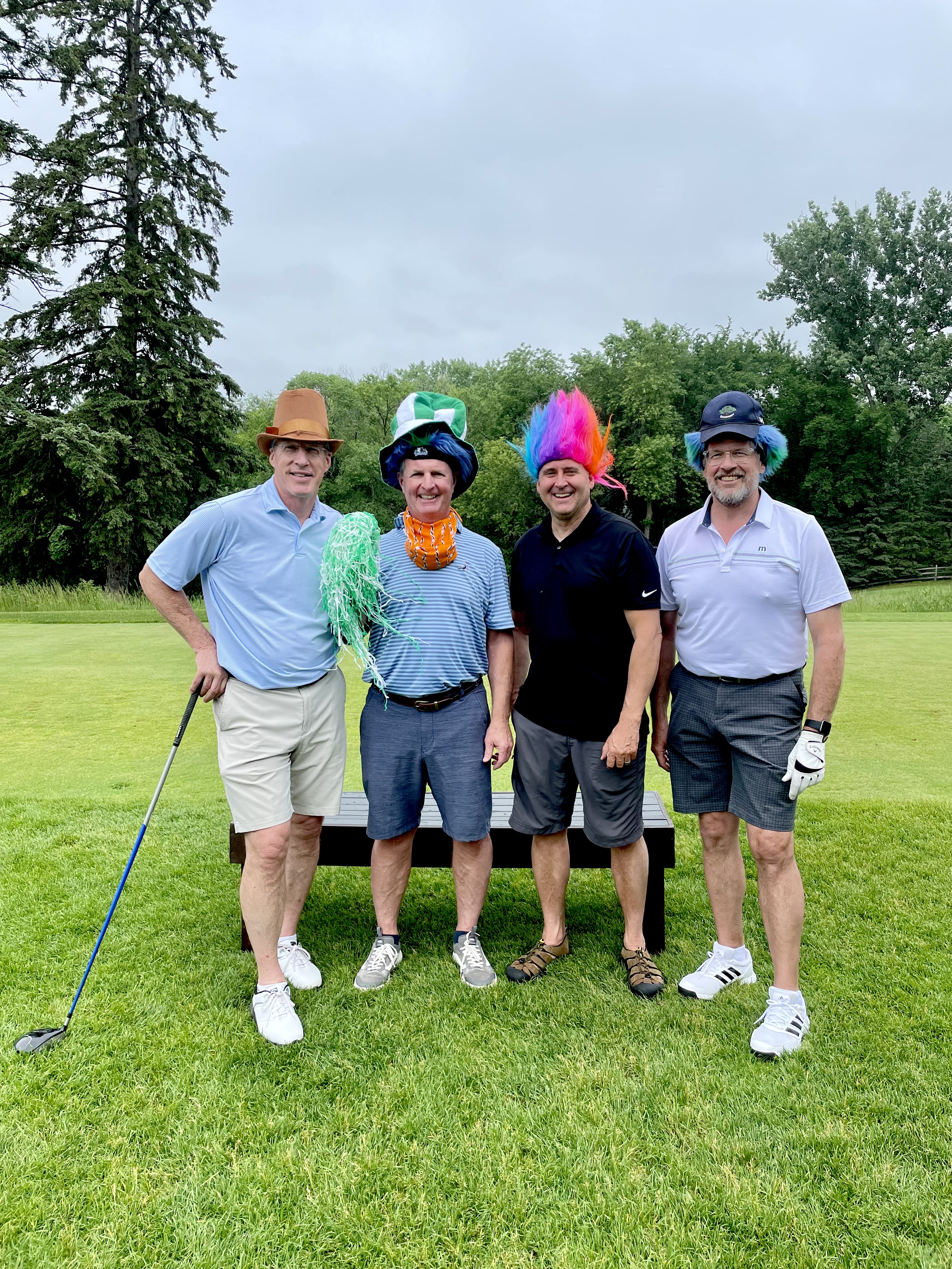 A group of four golfers pose for a photo while wearing wacky costumes and wigs on a golf course.