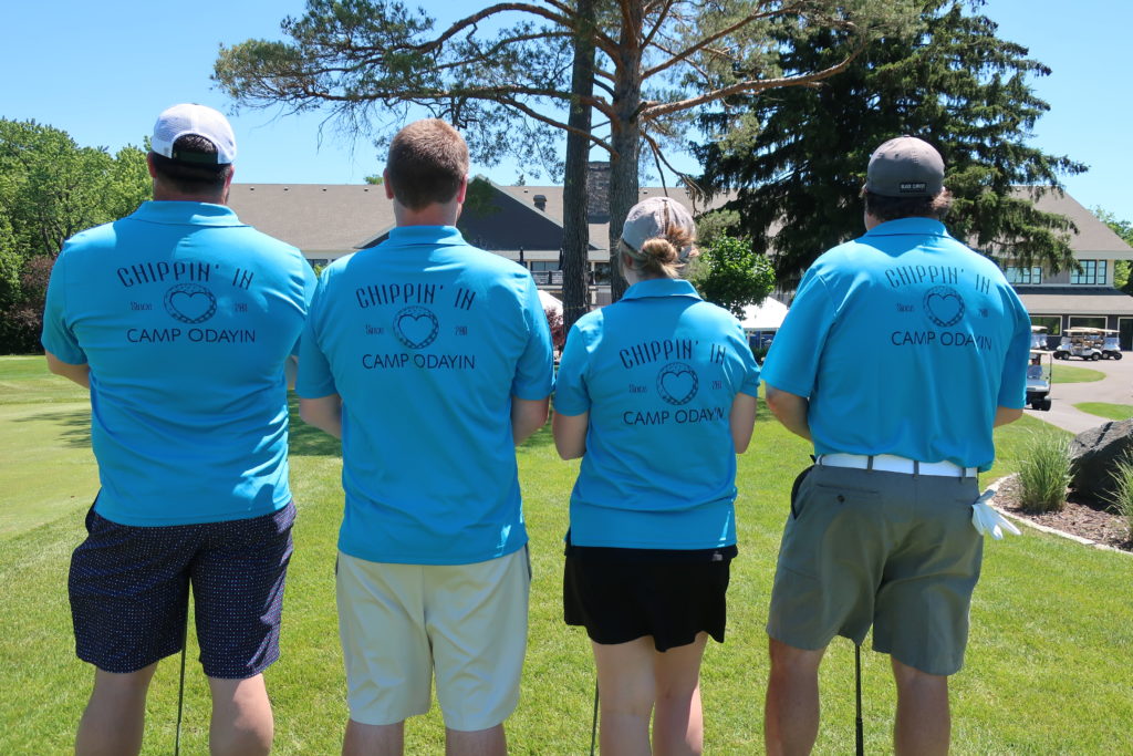A group of golfers with their backs towards the camera wearing matching polo shirts. The shirts read "Chippin' In" and "Camp Odayin".