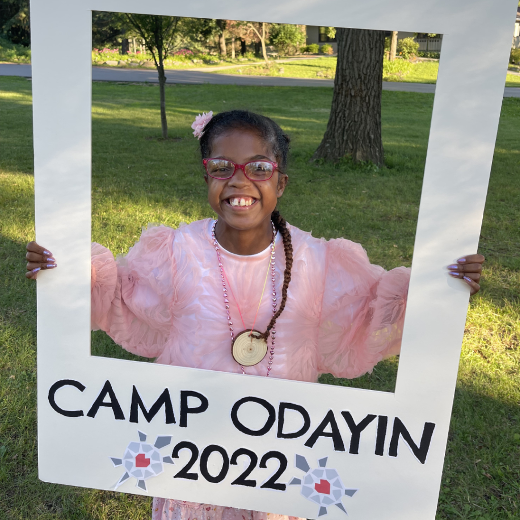 A camper in a fluffy pink shirt with a big smile holds up a picture frame that has the words "Camp Odayin 2022" written on it.