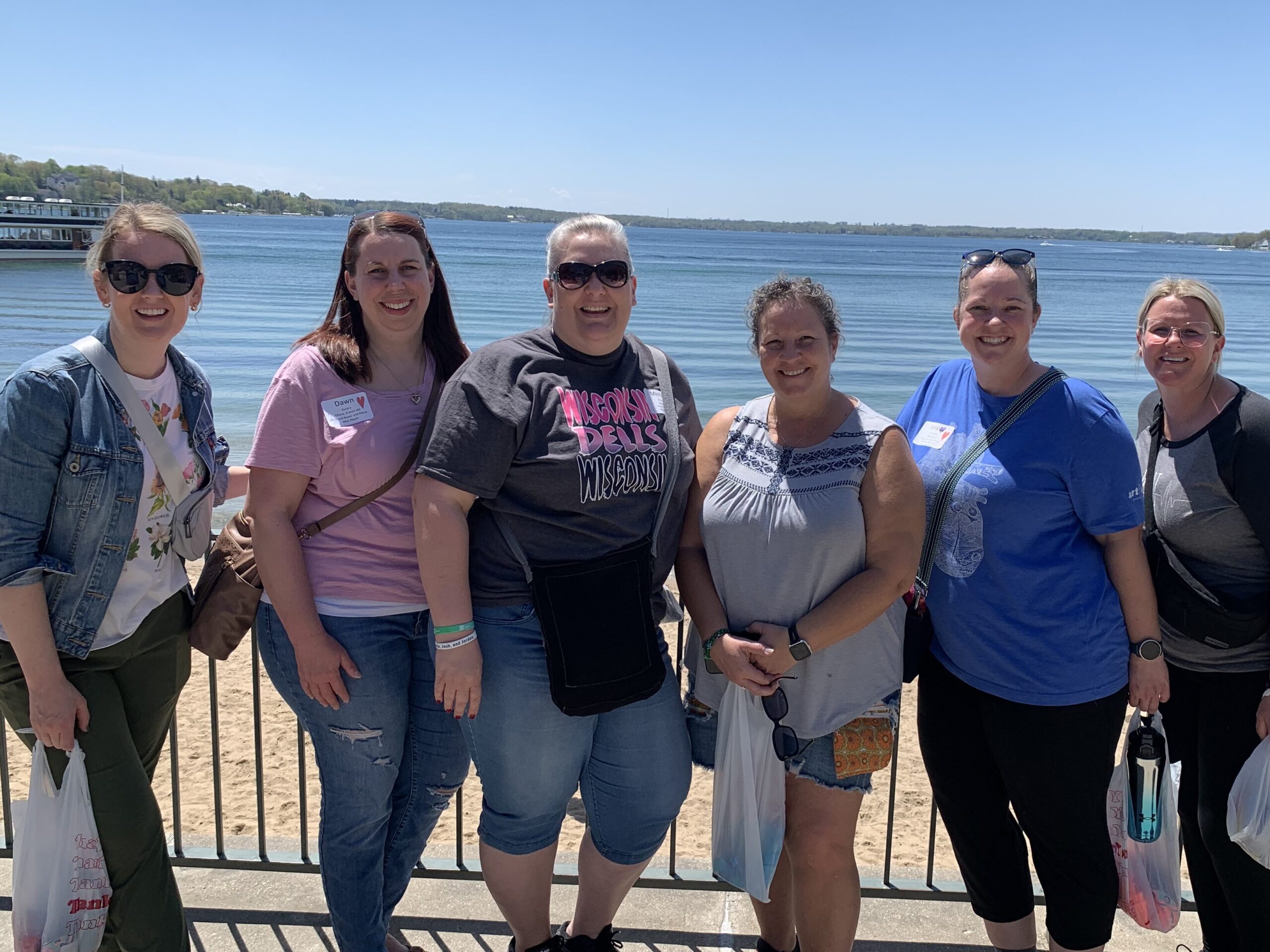 A group of six heart moms smile for a photo on a sunny day along the shores of Lake Geneva