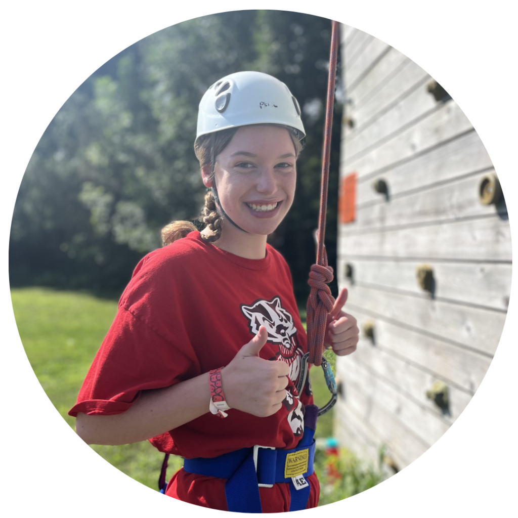 Camper giving a thumbs up while wearing a helmet and standing next to a climbing wall.
