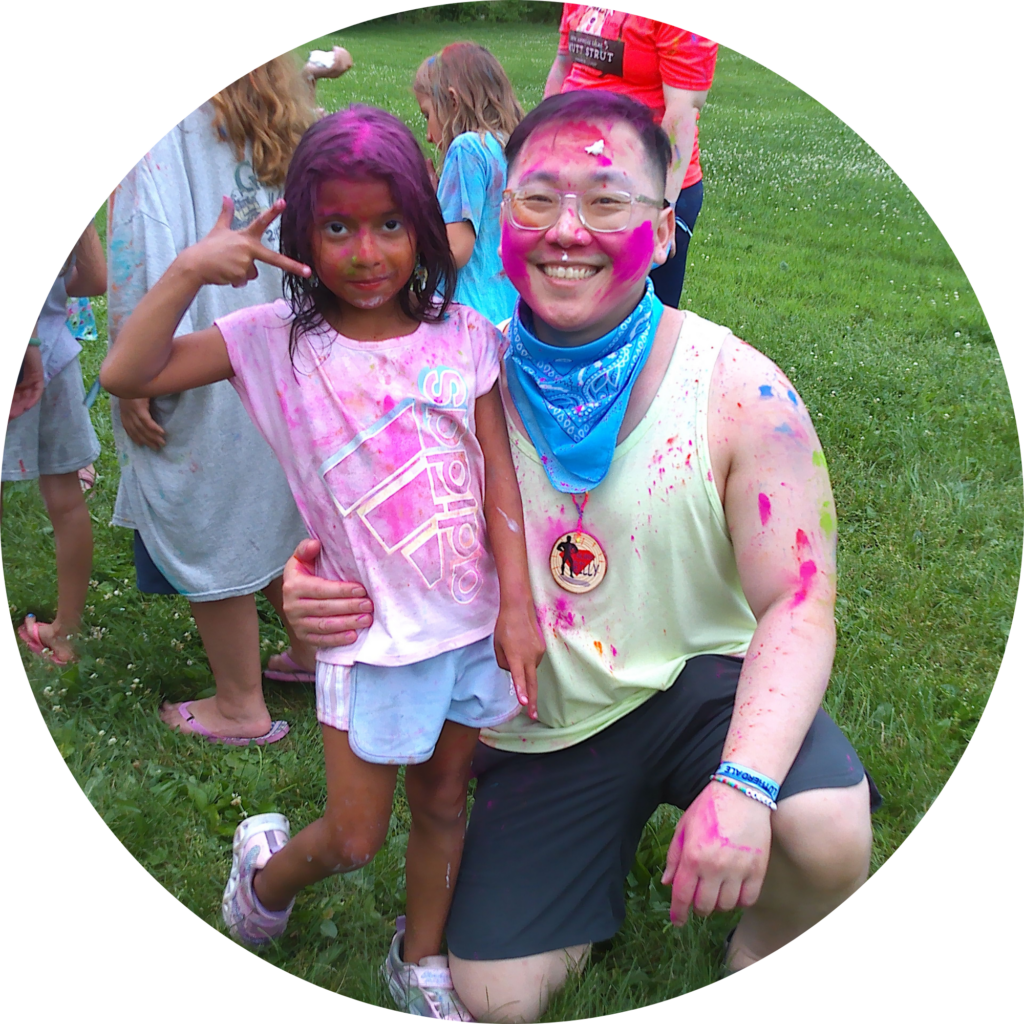A volunteer and a camper smile with their clothes and faces splattered with paint