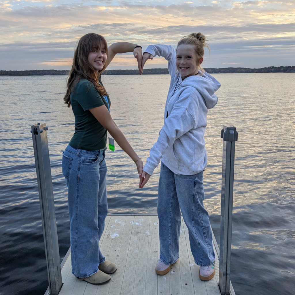 Two campers on a lake dock create a heart with their arms
